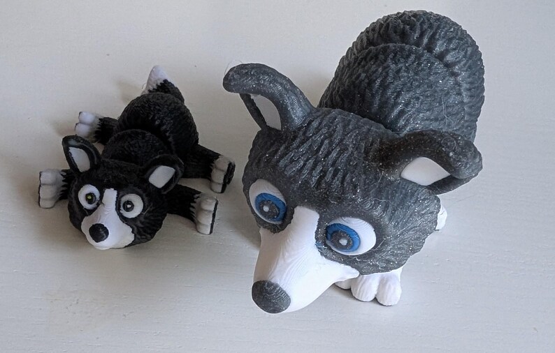 May include: Two husky dog figurines. One is lying down, the other is upright. Both have black and white markings, with blue eyes. The figurines are made of a textured material.
