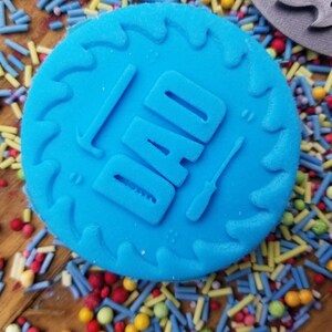 May include: A bright blue, circular cookie cutter with the word "DAD" embossed, along with a hammer and screwdriver. The cutter has a saw-tooth edge. Colorful sprinkles surround the cutter, suggesting a baking theme.