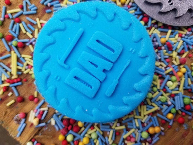 May include: A bright blue, circular cookie cutter with the word "DAD" embossed, along with a hammer and screwdriver. The cutter has a saw-tooth edge. Colorful sprinkles surround the cutter, suggesting a baking theme.