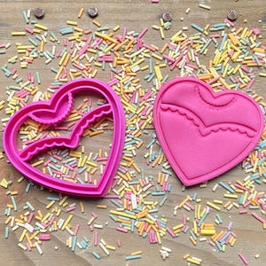 May include: Two pink heart-shaped cookie cutters, one with a scalloped edge and the other with a more intricate design. The cutters are on a wooden surface with colorful sprinkles.