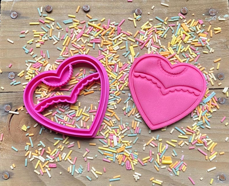 May include: Two pink heart-shaped cookie cutters, one with a scalloped edge and the other with a more intricate design. The cutters are on a wooden surface with colorful sprinkles.
