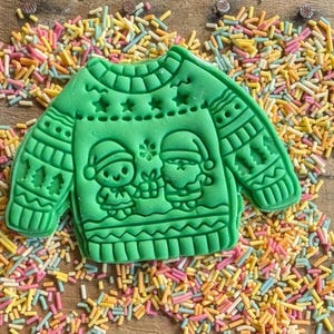 K&ouml;nnte beinhalten: Ein gr&uuml;ner Ausstecher in Form eines Weihnachtspullovers, verziert mit festlichen Mustern und zwei Cartoonfiguren mit Weihnachtsm&uuml;tzen. Der Ausstecher ist von bunten Streuseln umgeben und schafft eine fr&ouml;hliche Urlaubsszene.
