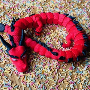 May include: A red and black segmented dragon toy with a textured surface. The dragon is curled up on a surface covered in colorful sprinkles.