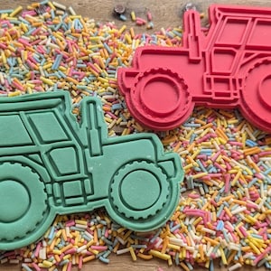 May include: Two tractor-shaped cookie cutters, one red and one green, on a bed of colourful sprinkles. The cutters have detailed wheels, cabs, and exhaust pipes. The sprinkles are a mix of pastel colours.