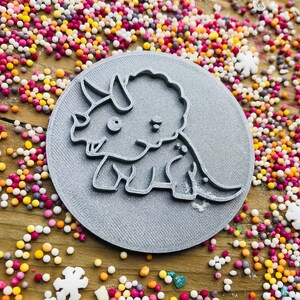 May include: A gray, circular cookie cutter featuring a cartoon triceratops dinosaur design. The cutter is surrounded by colorful sprinkles on a wooden surface, creating a playful and whimsical scene. Ideal for baking themed treats.