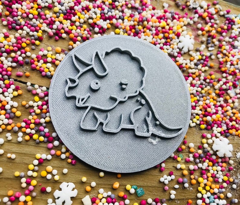 May include: A gray, circular cookie cutter featuring a cartoon triceratops dinosaur design. The cutter is surrounded by colorful sprinkles on a wooden surface, creating a playful and whimsical scene. Ideal for baking themed treats.