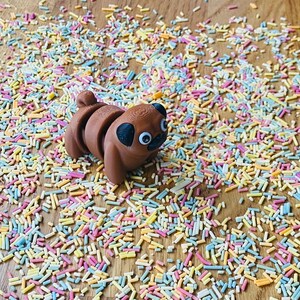Op de afbeelding: Een bruin mopsbeeldje met goggle-ogen dat op een houten oppervlak ligt bedekt met kleurrijke sprinkels.