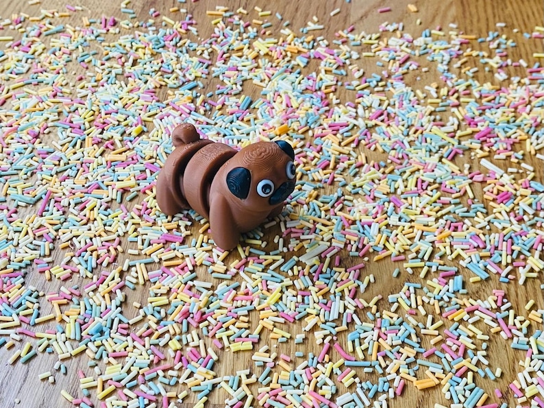Op de afbeelding: Een bruin mopsbeeldje met goggle-ogen dat op een houten oppervlak ligt bedekt met kleurrijke sprinkels.