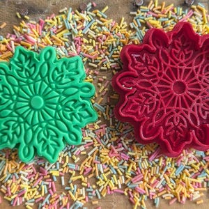 May include: Two snowflake-shaped cookie cutters, one green and one red, sit on a wooden surface covered in colorful sprinkles. The cutters have intricate floral designs, perfect for holiday baking.