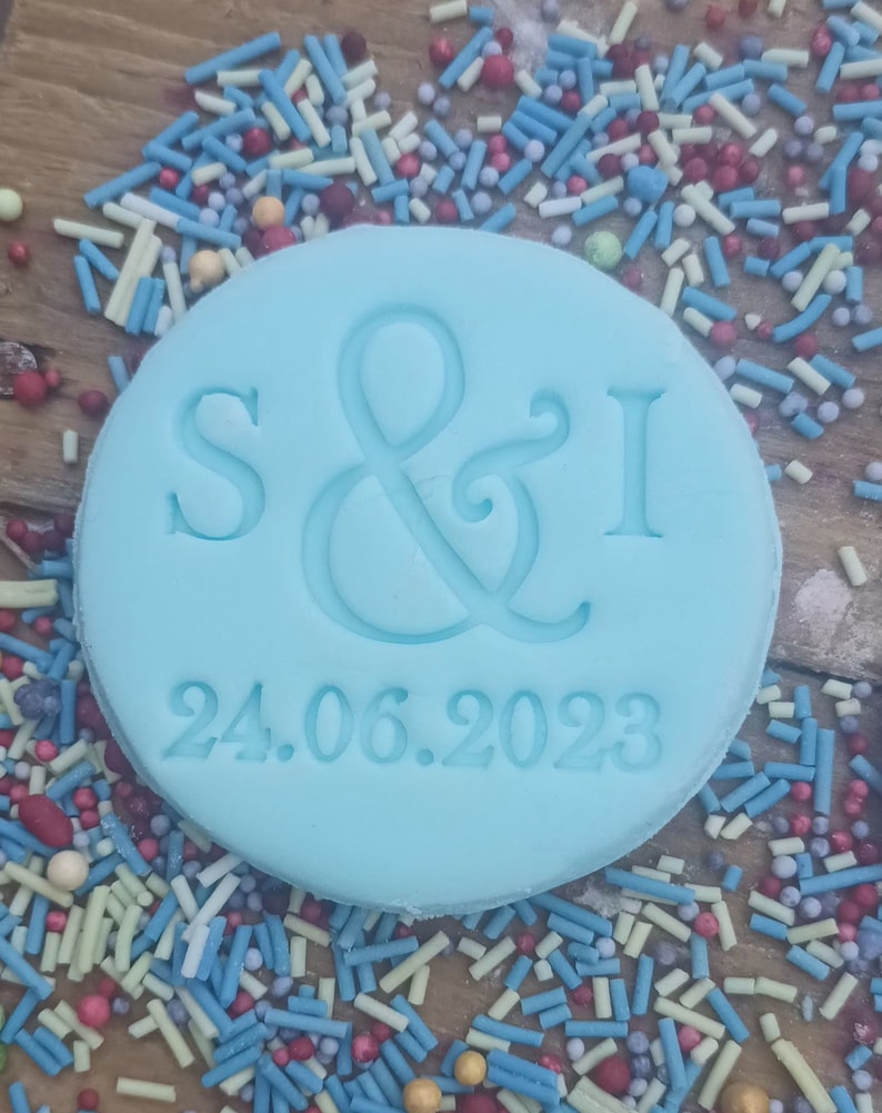 May include: A light blue round cookie with the letters "S&I" and the date "24.06.2023" embossed on it. The cookie is surrounded by colorful sprinkles.