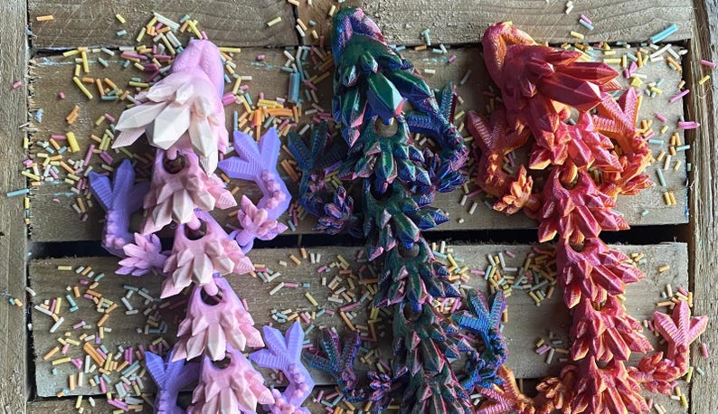 May include: Three 3D printed dragon figurines in pink, blue, and orange. The dragons are made of a textured material and have a spiky design.