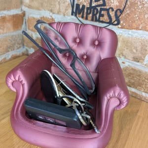 May include: A small, mauve-colored decorative chair-shaped tray. The tray holds a pair of eyeglasses, a car key fob, and a set of keys. The chair has a tufted back and armrests. The words "PRINT 2 IMPRESS" are visible in the background.