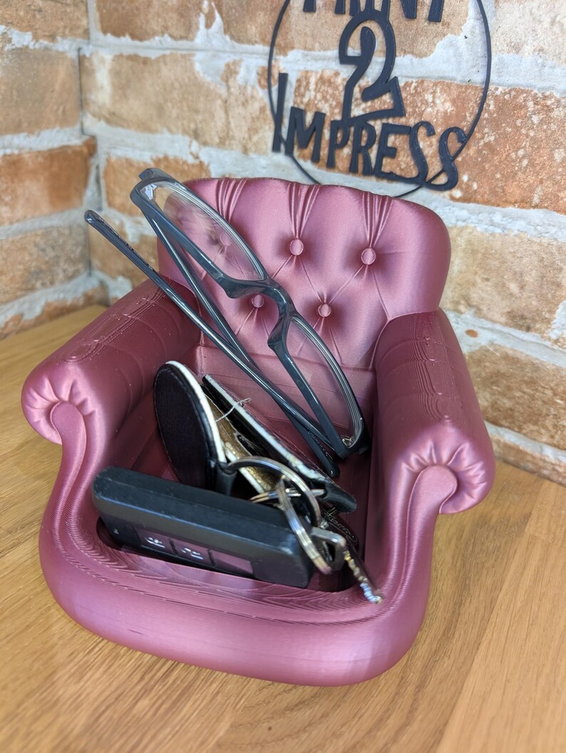 May include: A small, mauve-colored decorative chair-shaped tray. The tray holds a pair of eyeglasses, a car key fob, and a set of keys. The chair has a tufted back and armrests. The words "PRINT 2 IMPRESS" are visible in the background.