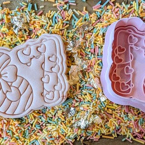 May include: Pink cookie cutter with a bow and heart design, next to a cut-out cookie. The cookie cutter and cookie are on a bed of colorful sprinkles and gold leaf flakes. Baking and crafting supplies.