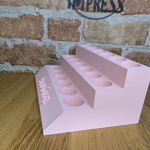 May include: A three-tiered, light pink organizer with multiple circular holes, likely for storing items. The word "IMPRESS" is visible in the background. The organizer is made of a solid material and sits on a wooden surface.