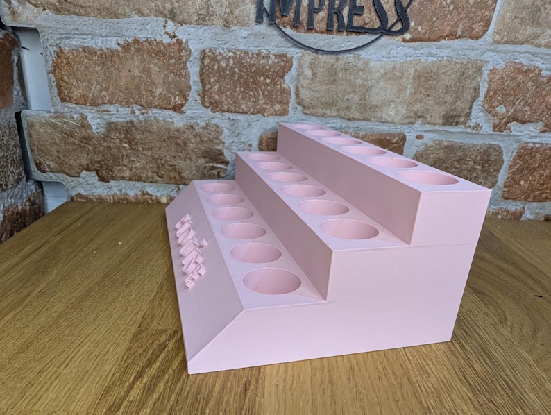 May include: A three-tiered, light pink organizer with multiple circular holes, likely for storing items. The word "IMPRESS" is visible in the background. The organizer is made of a solid material and sits on a wooden surface.