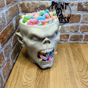 May include: A beige skull-shaped container filled with colorful gummy candies. The container has a wide open mouth with candies spilling out. The candies are in various colors, including pink, yellow, blue, and green.