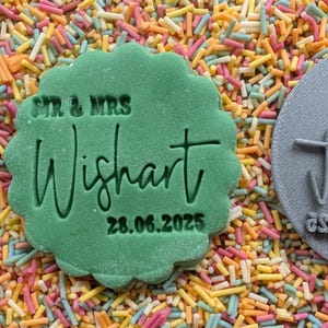 May include: A green, scalloped-edge cookie stamp with the text "SIR & MRS Wishart 28.06.2025" is surrounded by colorful sprinkles. A gray, circular stamp with text is also visible.