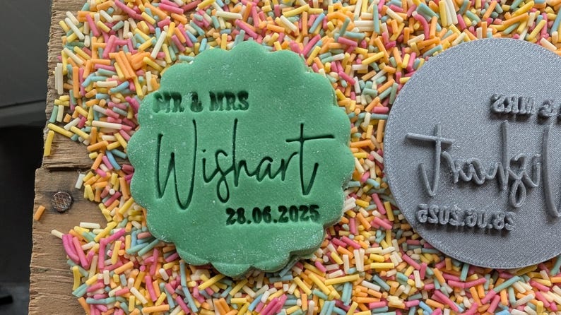 May include: A green, scalloped-edge cookie stamp with the text "SIR & MRS Wishart 28.06.2025" is surrounded by colorful sprinkles. A gray, circular stamp with text is also visible.