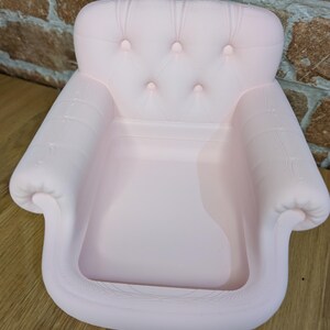 May include: A light pink miniature armchair with a tufted back and armrests. The small chair has a classic design with button detailing and a recessed seat. The chair is made of a solid material and is designed to be a decorative item.