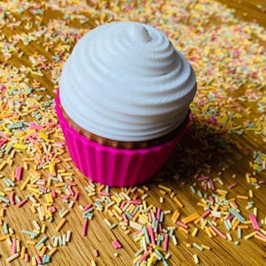 May include: A decorative cupcake with a white, swirled frosting top and a bright pink base. The cupcake sits on a wooden surface scattered with colorful sprinkles, creating a festive and playful scene. This item is perfect for home decor.