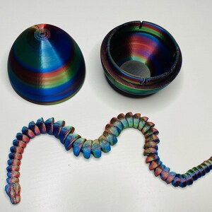 May include: A 3D printed iridescent blue, green, and red egg with a matching iridescent blue, green, and red snake. The egg is hollow and has a small hole in the top. The snake is segmented and has a curled tail.
