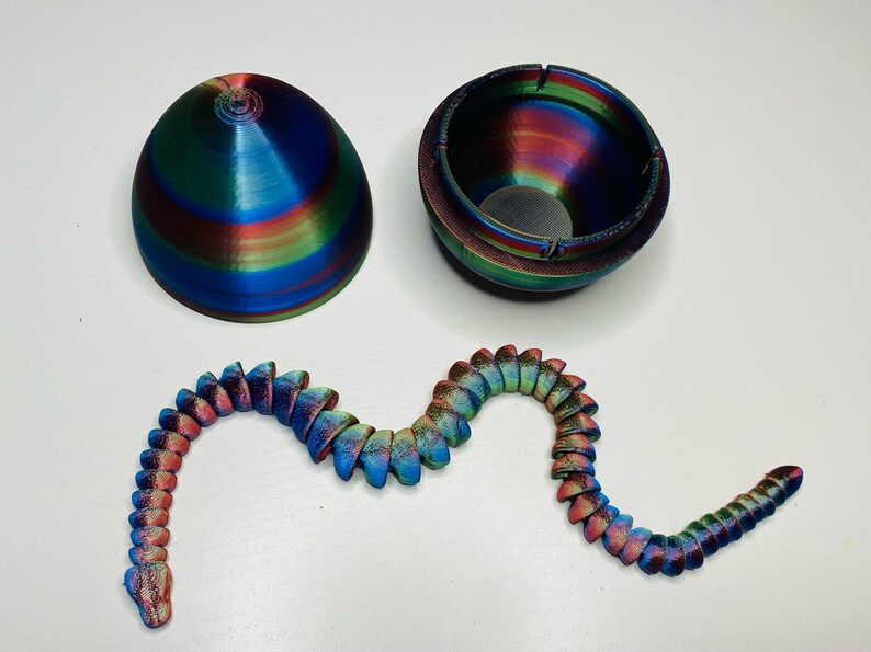 May include: A 3D printed iridescent blue, green, and red egg with a matching iridescent blue, green, and red snake. The egg is hollow and has a small hole in the top. The snake is segmented and has a curled tail.
