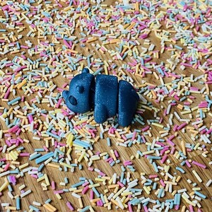 May include: A small, dark blue, 3D printed animal figurine shaped like a bear lying on its side. The figurine is surrounded by colorful sprinkles on a wooden surface.
