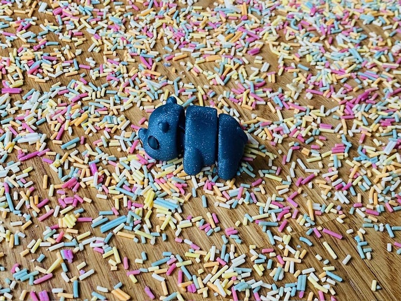 May include: A small, dark blue, 3D printed animal figurine shaped like a bear lying on its side. The figurine is surrounded by colorful sprinkles on a wooden surface.