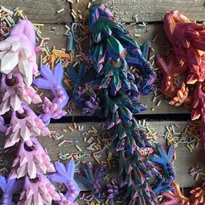 May include: Three 3D printed dragon figurines in pink, blue, and orange. The dragons are made of a textured material and have a spiky design.