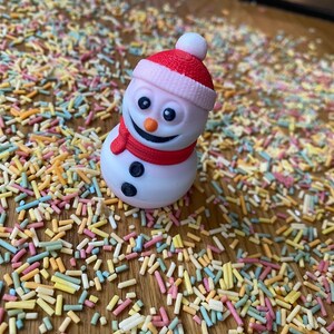 May include: A white snowman figurine with a red hat and scarf, sitting on a surface covered in colorful sprinkles.