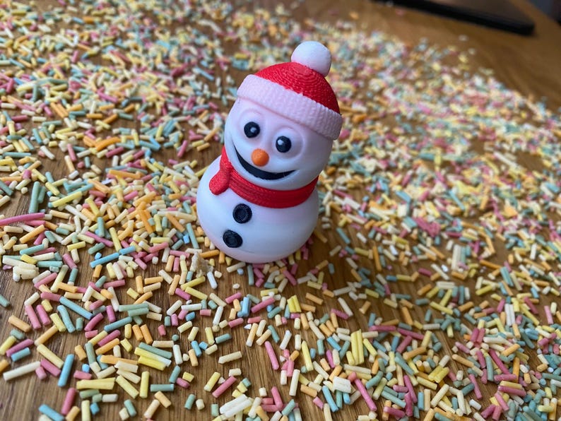 May include: A white snowman figurine with a red hat and scarf, sitting on a surface covered in colorful sprinkles.