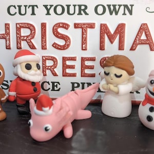 May include: A collection of Christmas-themed figurines, including a gingerbread man, Santa Claus, a pink axolotl, an angel, and a snowman. The figurines are in front of a sign that reads "CUT YOUR OWN CHRISTMAS TREES".