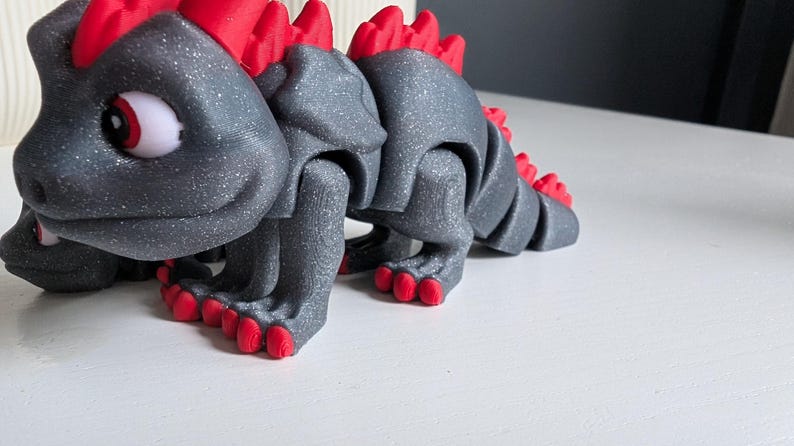 Op de afbeelding: Een grijze, beweegbare drakenfiguur met rode accenten. De draak heeft rode stekels op zijn rug en poten. De ogen zijn wit met rode pupillen. De figuur staat op een wit oppervlak.