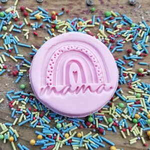 May include: A pink, circular cookie stamp with a rainbow design and the word "mama" in a script font. The stamp is surrounded by colorful sprinkles on a wooden surface.