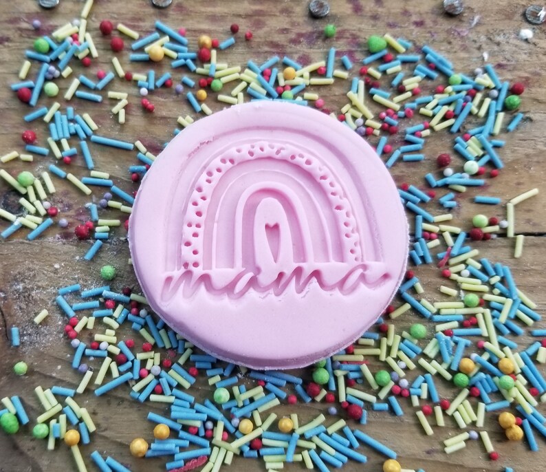 May include: A pink, circular cookie stamp with a rainbow design and the word "mama" in a script font. The stamp is surrounded by colorful sprinkles on a wooden surface.