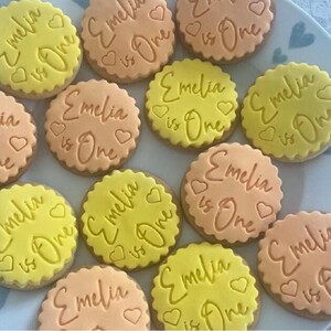 May include: A collection of decorated sugar cookies with yellow and orange icing. The cookies are decorated with the text "Emelia is One" in cursive writing.