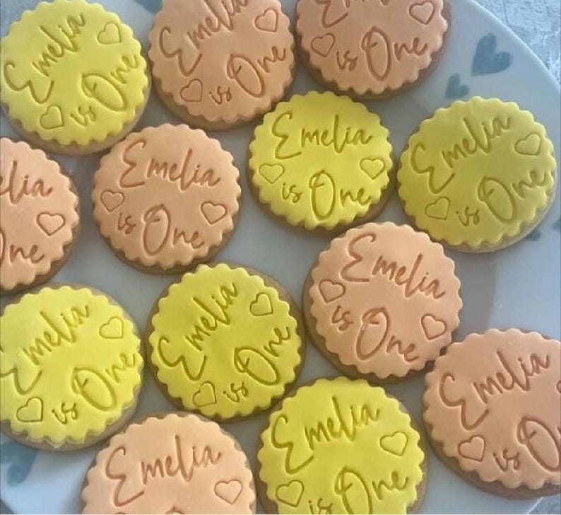 May include: A collection of decorated sugar cookies with yellow and orange icing. The cookies are decorated with the text "Emelia is One" in cursive writing.