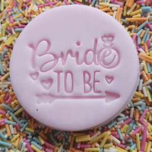 May include: A light pink, circular fondant with the words "Bride TO BE" embossed on it, along with a ring and arrow design. The fondant is surrounded by colorful sprinkles, creating a festive and celebratory look.