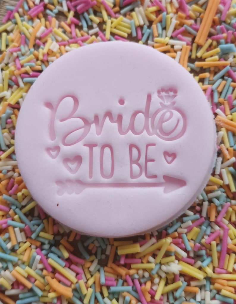 May include: A light pink, circular fondant with the words "Bride TO BE" embossed on it, along with a ring and arrow design. The fondant is surrounded by colorful sprinkles, creating a festive and celebratory look.