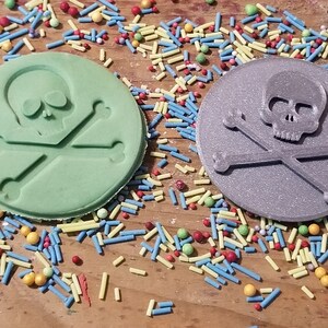 May include: Two round, skull and crossbones-shaped objects. One is light green, the other is gray. Both have the skull and crossbones design embossed on the surface. Colorful sprinkles are scattered around the objects on a wooden surface.