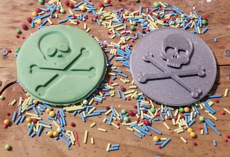 May include: Two round, skull and crossbones-shaped objects. One is light green, the other is gray. Both have the skull and crossbones design embossed on the surface. Colorful sprinkles are scattered around the objects on a wooden surface.
