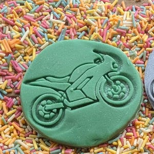 May include: A green, circular cookie cutter with a motorcycle design. The motorcycle is depicted in a side profile, with detailed wheels and body. The cookie cutter is surrounded by colorful sprinkles.