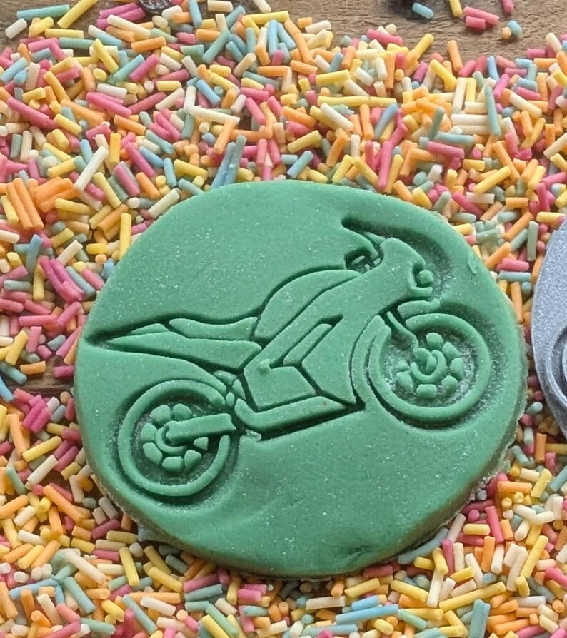 May include: A green, circular cookie cutter with a motorcycle design. The motorcycle is depicted in a side profile, with detailed wheels and body. The cookie cutter is surrounded by colorful sprinkles.