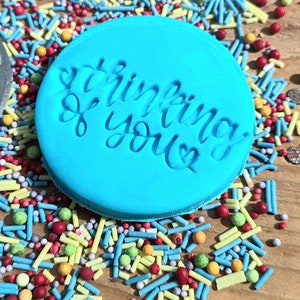 May include: A round, light blue object with the words "thinking of you" embossed in a cursive font. The object is surrounded by colorful sprinkles in various shapes and sizes, including red, yellow, green, and blue.