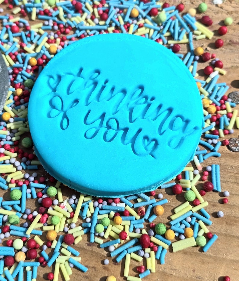 May include: A round, light blue object with the words "thinking of you" embossed in a cursive font. The object is surrounded by colorful sprinkles in various shapes and sizes, including red, yellow, green, and blue.