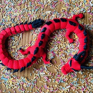 May include: A red and black dragon toy with a textured surface. The dragon is curled up in a circle and is surrounded by colorful sprinkles.