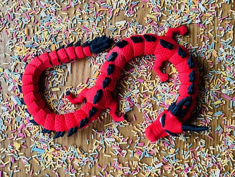 May include: A red and black dragon toy with a textured surface. The dragon is curled up in a circle and is surrounded by colorful sprinkles.