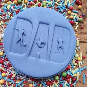 May include: A light blue, circular cake decoration with the word "DAD" embossed in large letters. The letters feature soccer player silhouettes and a soccer ball. The cake is surrounded by colorful sprinkles on a wooden surface.