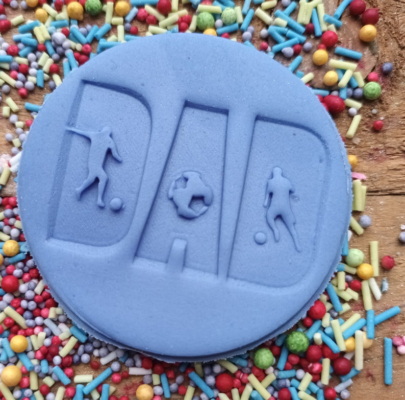 May include: A light blue, circular cake decoration with the word "DAD" embossed in large letters. The letters feature soccer player silhouettes and a soccer ball. The cake is surrounded by colorful sprinkles on a wooden surface.